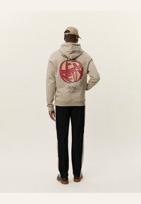 Sudadera beige con un gráfico de una manzana roja y texto en la parte posterior, combinada con pantalones negros que tienen una franja lateral blanca y zapatos marrones.