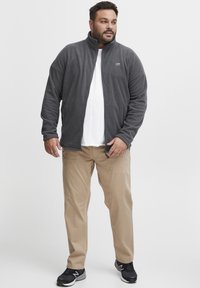 Veste en polaire grise avec fermeture éclair intégrale, accompagnée d'un pantalon beige clair. L'ensemble comprend un t-shirt blanc et des chaussures de sport noires.