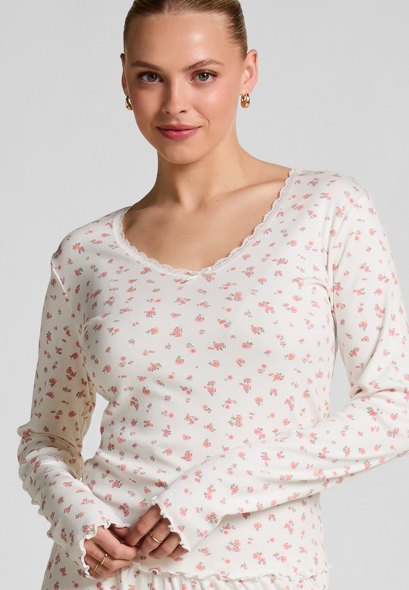Long-sleeve ribbed top in cream with a floral pattern in pink and green, featuring a scalloped V-neck and ruffled cuffs.