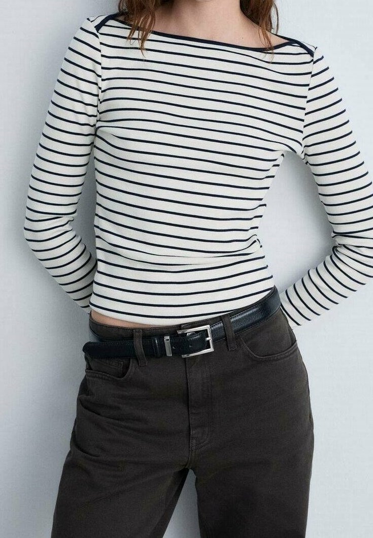 Femme portant un haut blanc à manches longues avec des rayures horizontales noires et un pantalon foncé avec une ceinture noire, debout sur un fond uni.