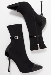 Botas altas de tacón negro con parte superior de tejido suave, puntera de charol, cremallera lateral y detalle decorativo de hebilla en el tobillo.