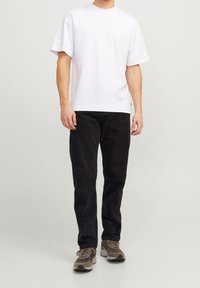 White cotton t-shirt with a relaxed fit, paired with black corduroy pants. Worn with grey and beige sneakers. Plain background.