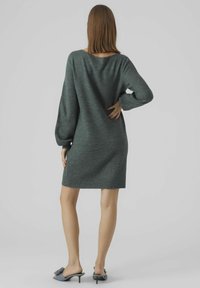 Abito maglione over-size lungo fino al ginocchio in maglia verde scuro. Presenta maniche lunghe, un ampio scollo e polsini a coste, con una texture morbida.