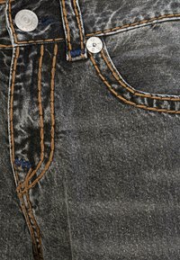 Dark gray denim jeans with visible stitching in brown and blue, featuring metal buttons and a pocket detail, showing a textured fabric surface.