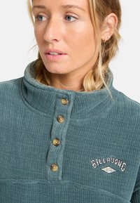 Suéter de canalé en color teal con cuello alto, que presenta cuatro botones en tono marrón claro y un logo bordado en el pecho. Textura suave y diseño estructurado.