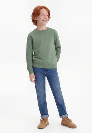 Αγόρι με κόκκινα μαλλιά στέκεται, φοράει πράσινο μπλουζάκι με μακριά μανίκια, μπλε τζιν με γυρισμένα μπατζάκια και ανοιχτοκαφέ μπότες με κορδόνια, χαμογελά και κοιτάζει δεξιά.