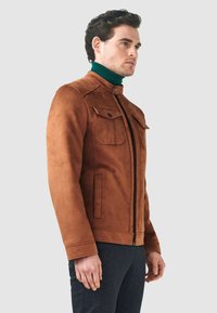 Brun mocka jacka med hög krage, två bröstfickor och sidfickor. Har en frontdragkedja och strukturerade axlar.