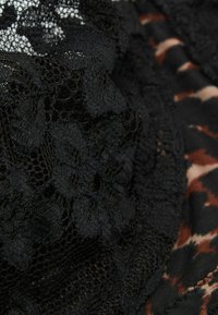 Černá krajka se květinovými vzory, s otevřenou síťovinou a pevným materiálem, vrstvená přes vzorované pozadí v odstínech hnědé a béžové.