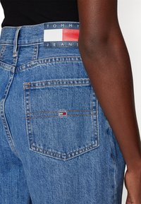Jeans de mezclilla azul con una cintura alta, que cuentan con bolsillos traseros y una etiqueta de logo de Tommy Jeans en rojo, blanco y azul en la cinturilla.