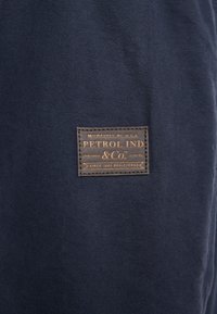 Navyblauwe textiel met een rechthoekig bruin leren label waarop "PETROL IND & Co." is gestikt.