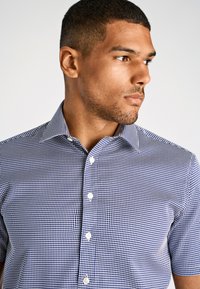 Camicia a maniche corte con bottoni, in pattern a quadretti blu navy e bianchi, tessuto leggero, colletto strutturato e cinque bottoni sulla parte frontale.