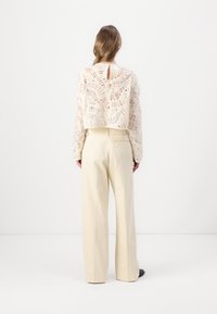 Häkeltop in Beige mit Struktur und langen Ärmeln, kombiniert mit hochtaillierten, hellbeigen weit geschnittenen Hosen. Minimalistisches Design mit einem Schlüsselloch im Rücken.