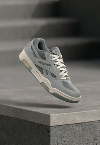 Svijetlosive i bijele Reebok tenisice s mrežastim i od nubuka panelima lebde iznad betonskog ruba.