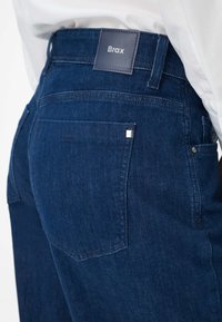 Donkerblauwe denim jeans met een gladde textuur, voorzien van een vierkante metalen accenten op de achterzak en een leren merklabel op de tailleband.