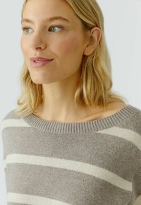 Blonde vrouw met groene ogen, gekleed in een beige en witte gestreepte gebreide trui, kijkt iets naar links tegen een neutrale lichte achtergrond.