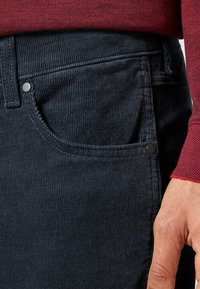 Marineblauwe corduroy broek met een gestructureerde afwerking, voorzien van een zijzak en een zwarte metalen knoopaccent. Rood sweater zichtbaar erboven.