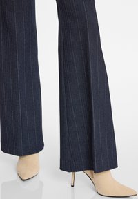 Dunkelblaue rot gestreifte weit geschnittene Hose in Kombination mit beigefarbenen Wildleder-Pumps mit spitzer Zehenform auf einer reflektierenden weißen Oberfläche.