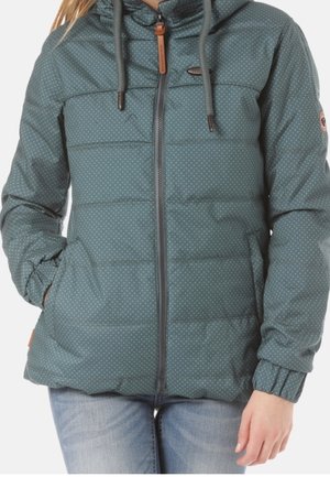 Kvinde iført en teal dunjakke med hætte, lille prikmønster, frontlynlås og elastiske manchetter, kombineret med lyseblå jeans.