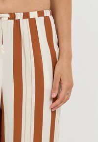 Pantaloni a strisce crema e ruggine con un tessuto morbido, vita elastica e dettaglio con cordino. Le strisce verticali amplificano la lunghezza visiva.