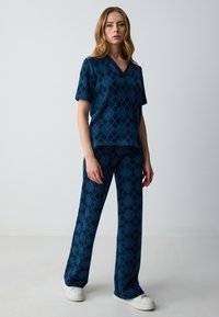 Completo in maglia a motivo argyle blu e verde. La parte superiore presenta un collo a V e maniche corte, mentre i pantaloni sono a gamba larga. Scarpe bianche.