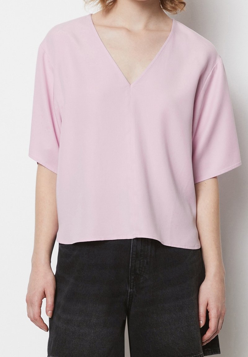 Femme portant un chemisier rose clair à manches courtes et col en V, et un short en jean noir taille haute, debout devant un fond uni.