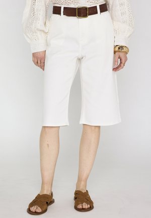 Femme portant un short blanc mi-cuisses, une ceinture en cuir marron, un chemisier blanc ajouré, un bracelet en or et des sandales marron, debout devant un fond uni.