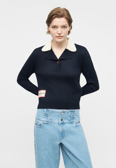 Γυναίκα που φοράει μπλε πουλόβερ με λευκό γιακά sherpa και ανοιχτό μπλε ψηλόμεσο τζιν, στέκεται με τα χέρια πίσω από την πλάτη σε απλό φόντο.