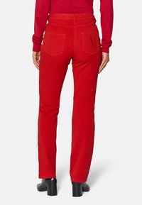 Rote Baumwollhose mit geradem Bein, ausgestattet mit zwei Gesäßtaschen und einem gestickten "M"-Detail. Kombiniert mit einem taillierten roten Oberteil und hochhackigen Schuhen.