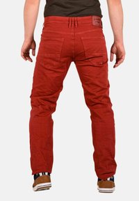 Pantalones de algodón rojos con cinco bolsillos, pasadores para cinturón y un parche con logo en la parte trasera. Presenta un ajuste slim con una textura ligeramente arrugada.