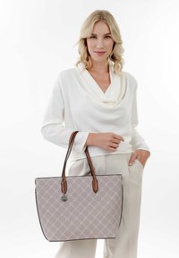 Sac fourre-tout gris à motif avec des bords noirs et des poignées marron, orné d'une étiquette décorative. Le modèle porte une blouse blanche et un pantalon clair.