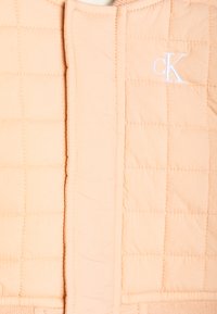 Lys fersken fôret vest med firkantet quilting, en glatt tekstur og den kontrasterende hvite "CK" logoen sydd foran.