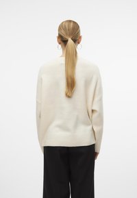 Cremefarbenes Strickpullover mit lockerer Passform, überschnittenen Schultern und geripptem Saum. Kombiniert mit schwarzen Hosen, die eine glatte Textur zeigen.