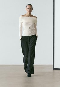 Kremowy top z odkrytymi ramionami i długimi rękawami w połączeniu z czarnymi spodniami z wysokim stanem. Gładka faktura, minimalistyczny krój i dopasowana sylwetka.