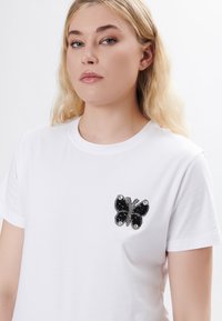 Hvid bomulds t-shirt med en sort juvelbesat sommerfugle-appliqué på venstre bryst, med rund hals og korte ærmer.