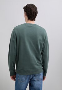 Felpa verde con maniche lunghe, collo a giro in coste e tessuto strutturato. Indossata con jeans azzurri, presenta una vestibilità comoda e un design dell'orlo.