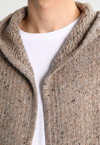Cárdigan de punto en tonos beige y marrón, con un gran cuello, textura con fibras salpicadas y diseño de frente abierto sobre una camisa blanca.