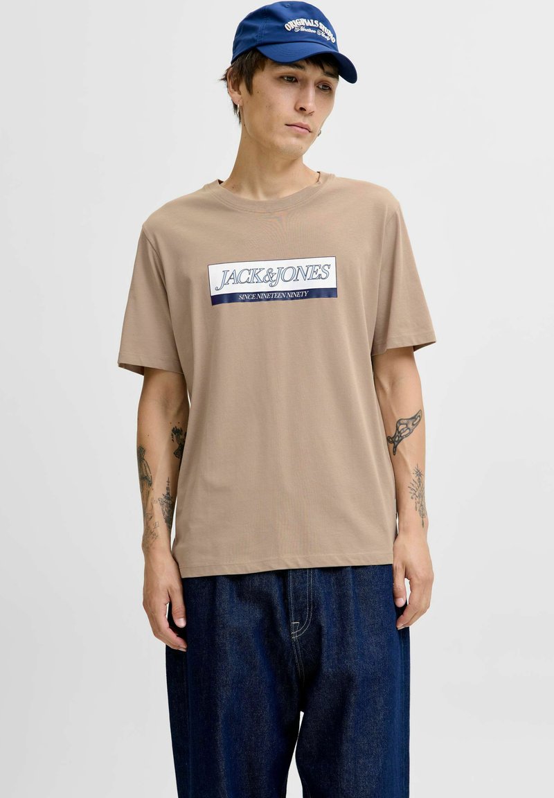 Beige T-shirt med korte ærmer med et rektangulært navy og hvidt "JACK&JONES" grafiklogo. Paret med løstsiddende mørkeblå denim jeans.
