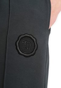 Black athletic pants featuring a textured fabric. A round, embossed black logo patch is on the left thigh. Stitching is subtle and tonal.