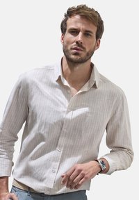 Helles beige Button-Down-Hemd mit vertikalen weißen Streifen, langen Ärmeln und klassischem Kragen. Kombiniert mit einer einfachen Uhr und hellen Hosen.