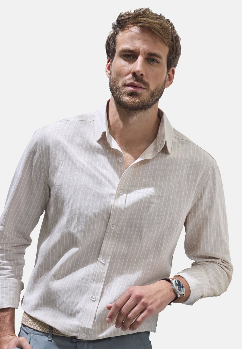 Helles beige Button-Down-Hemd mit vertikalen weißen Streifen, langen Ärmeln und klassischem Kragen. Kombiniert mit einer einfachen Uhr und hellen Hosen.