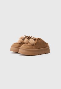 Pantofi slip-on din suede maro cu platformă, cu șireturi decorate cu trei pom-pom-uri pufoase pe fiecare pantof, așezați pe un fundal simplu.