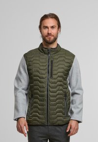 Chaleco tipo plumón en verde olivo con textura acolchada, mangas grises, cuello alto y bolsillos frontales con cremallera. Diseño ligero y moderno.