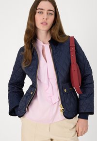 Navy quilted jacket with gold zipper and hardware, featuring a black collar. Worn over a pink ruffled blouse and beige trousers, with a red strap bag.