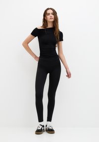 Fekete, testhezálló felső, egy vállon viselhető dizájnnal és redős textúrával, fekete leggings-szel párosítva. A modellen fekete tornacipő van fehér fűzőkkel.