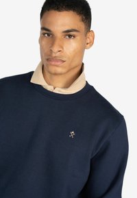 Sudadera de color azul marino con un collar beige, que presenta un pequeño logotipo metálico en el pecho, fabricada con una tela suave y ligeramente texturizada.