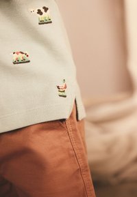 Polo vert menthe avec motifs brodés d'animaux de la ferme. Comprend des manches courtes, un col côtelé et des fentes latérales. Porté avec un pantalon marron.