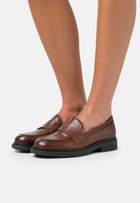 Mocassins en cuir marron avec un bout rond, des coutures décoratives et une semelle en caoutchouc noir contrastante. Texturé lisse, accents minimaux.