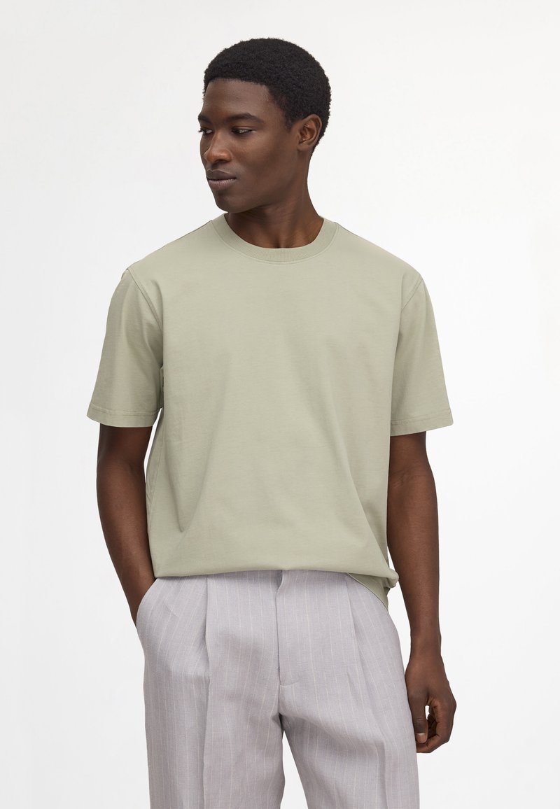 Jeune homme aux cheveux courts et bouclés porte une chemise vert clair à manches courtes et un pantalon gris clair à fines rayures, mains dans les poches, regardant vers la gauche.
