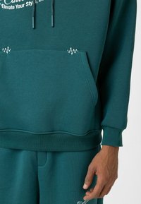 Tegengroene hoodie met een kangoeroezak, voorzien van witte stikdetails en een trekkoord met bijpassende uiteinden. Zachte, gestructureerde stof.