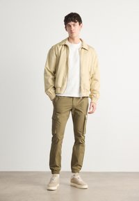 Lichtbeige bomberjack met elastische boorden, gecombineerd met olijfgroene cargobroeken met zijzakken en elastische enkels. Beigekleurige sneakers maken de look compleet.